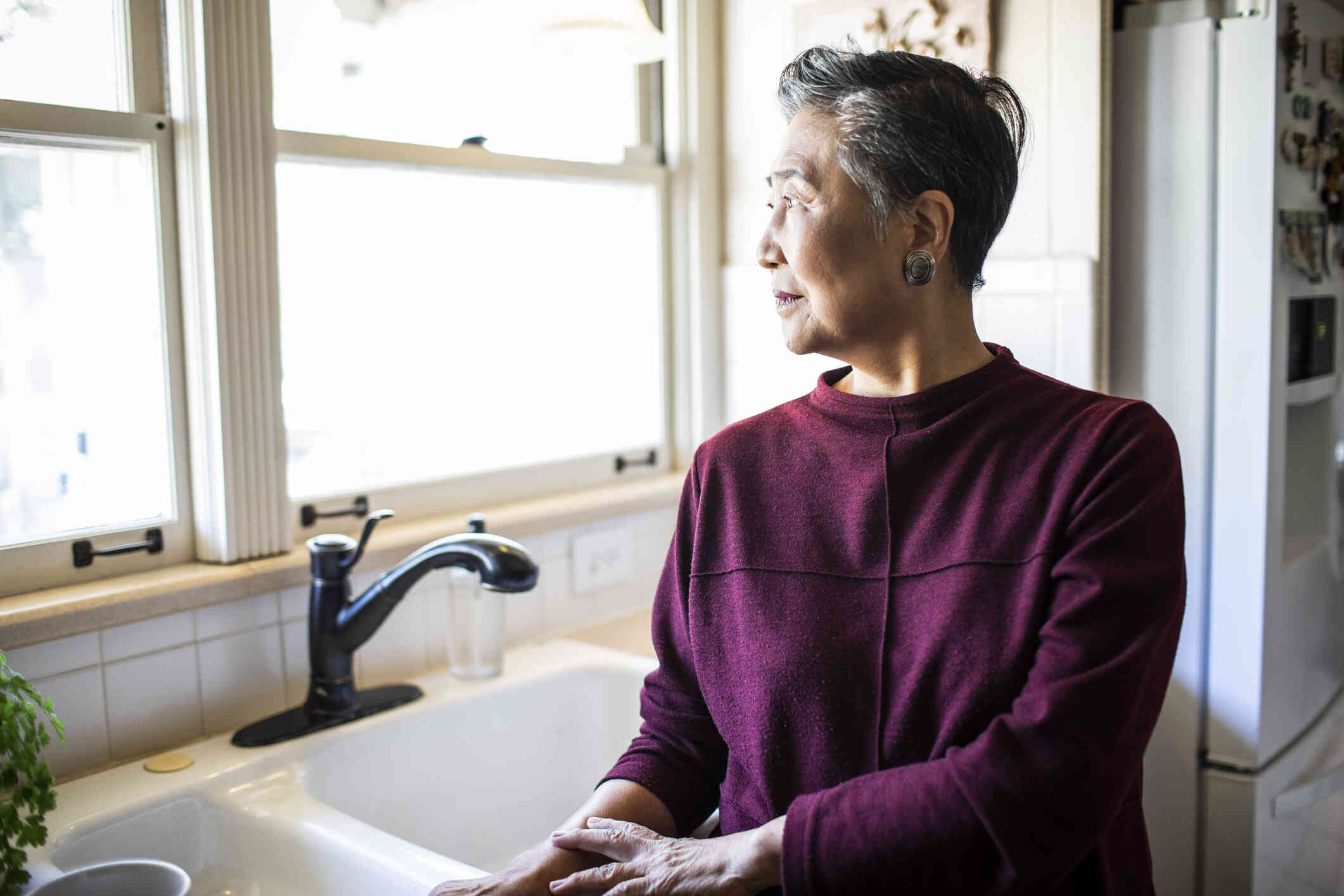 An elderly woman in a purple sweater stands in her kitchen and gazes out of the window while deep in thought.